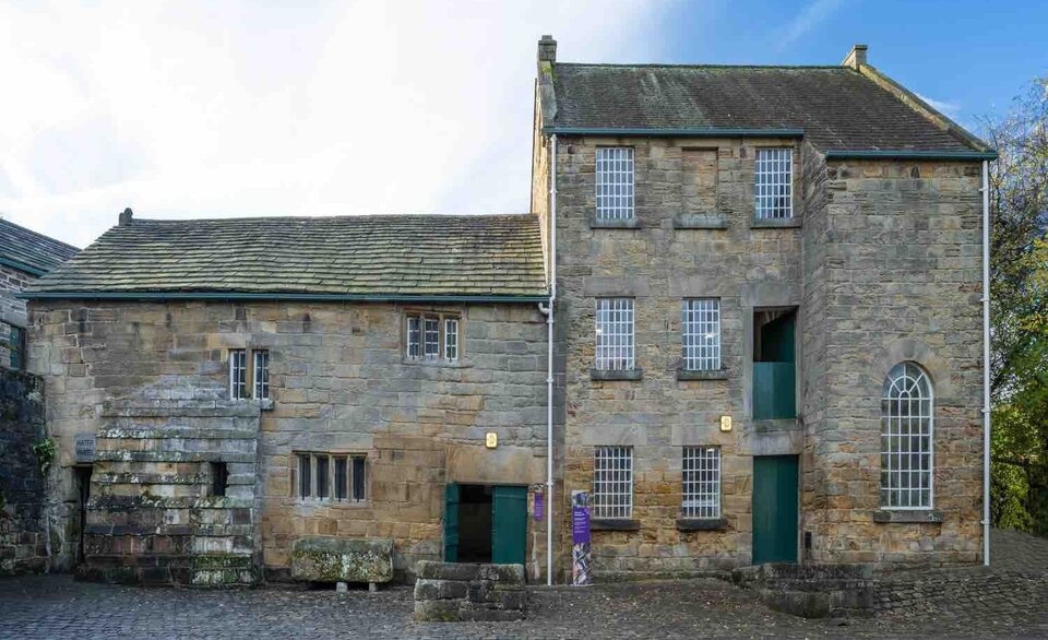 A historic stone building with two sections: a shorter wing with a sloped roof and small windows, and a taller three-storey section with larger barred windows and green doors.